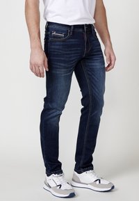 Jeans de mezclilla azul oscuro con un corte Slim, con costuras naranjas, bolsillos frontales y cremallera, combinados con zapatillas deportivas blancas y grises.