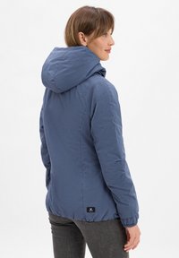 Veste à capuche bleue pour femmes avec une texture lisse et des poignets élastiques. Présente un logo au bas du dos et est conçue pour un look ajusté.