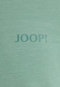 Tejido de un verde menta claro, con textura acanalada, que presenta la palabra "JOOP!" en un tono de verde más oscuro, en un estilo moderno y en negrita.