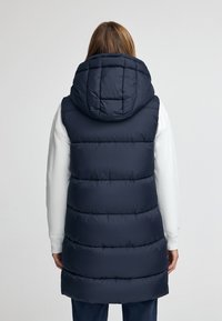 Mörkblå pufferväst med huva, med kviltad design, medellängd och en mjuk textur. Bärs över en ljusfärgad långärmad skjorta.