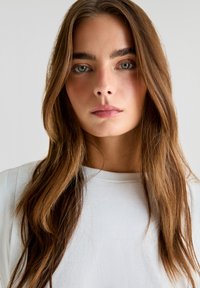 Hvid crew-neck top, glat stof, langt brunt hår med lysere striber, naturlig makeup, neutral baggrund. Fokus på ansigt og halsudskæring.