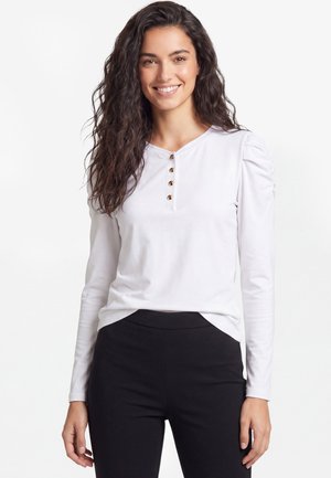 Ung kvinde med langt krøllet hår iført en hvid langærmet top med knapper og sorte bukser, smilende til kameraet.