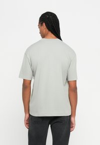 Άτομο με μακριά �πλεχτά μαλλιά γυρίζει πλάτη, φορώντας ένα ανοιχτό γκρι κοντομάνικο μπλουζάκι και σκούρα γκρι τζιν, μπροστά σε ένα λευκό φόντο.