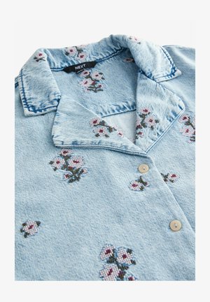 Chemise en denim bleu clair avec col, ornée de broderies florales en rose, blanc et vert. Deux boutons beiges à l'avant.
