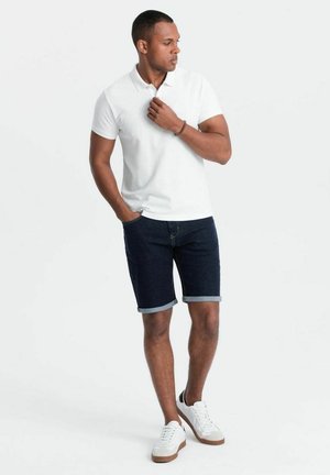 Camisa polo blanca combinada con pantalones cortos de mezclilla oscuros. Los pantalones cortos están doblados en el dobladillo. El modelo lleva zapatillas blancas con acentos marrones.