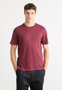 Jonge man in een kastanjebruine T-shirt met korte mouwen met het "HUGO" logo en zwarte broek, staand met zijn handen in zijn zakken tegen een witte achtergrond.