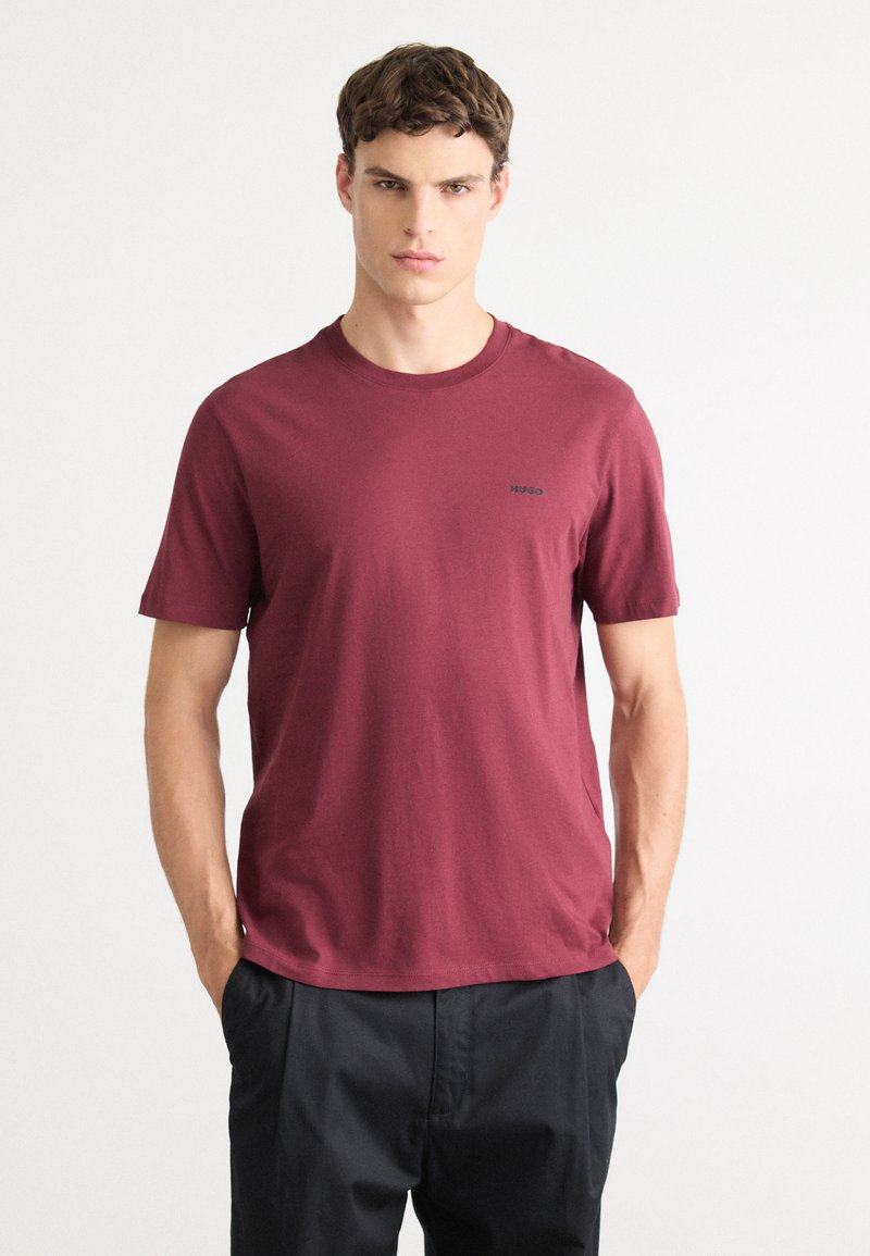 Jonge man in een kastanjebruine T-shirt met korte mouwen met het "HUGO" logo en zwarte broek, staand met zijn handen in zijn zakken tegen een witte achtergrond.