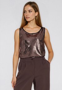 Kobieta ubrana w bezrękawnik z błyszczących brązowych cekinów, wpuszczony w wysoki stan ciemnych brązowych spodni, z jedną ręką w kieszeni.