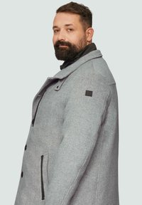 Grijze wollen jas van een wolblend met een notch-kraag, zwarte accentzak-ritsen en een logo patch op de schouder. Gestructureerd visgraatpatroon.