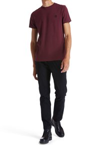 Burgundy cotton t-shirt with short sleeves and crew neckline; features a small black logo on the chest; paired with black pants and boots.