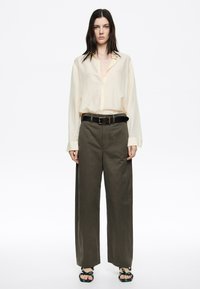 Camicia button-up beige chiaro con maniche lunghe, abbinata a pantaloni larghi verde oliva e una cintura nera. Le calzature presentano un design con cinturino testurizzato nero.