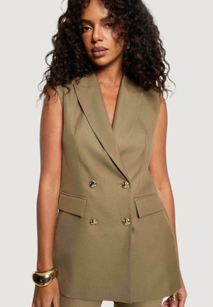 Donna con capelli ricci che indossa un blazer beige doppiopetto senza maniche con bottoni dorati e pantaloni abbinati, accessoriata con un braccialetto spesso d'oro.