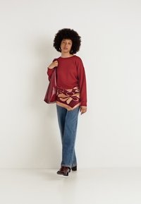 Felpa rossa a maniche lunghe, gonna stampata, jeans a palazzo blu, stivali marroni alla caviglia, porta una borsa rossa lucida. Sfondo semplice.