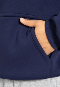 Sweat à capuche bleu marine avec une poche avant, en matériau coton doux, avec des poignets et un ourlet côtelés. Main reposant à l'intérieur de la poche. Tissu gris visible en dessous.