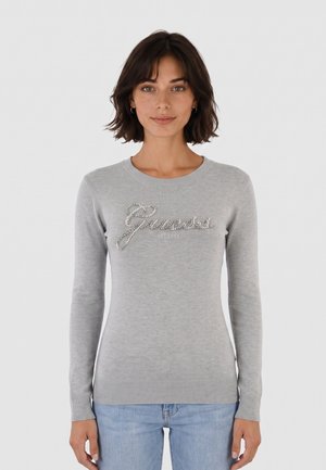 Mujer con cabello corto y oscuro que lleva un suéter gris claro con el logo "Guess" en pedrería y vaqueros azul claro, de pie frente a un fondo blanco.