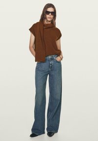 Top in maglia marrone con design drappeggiato, abbinato a jeans in denim azzurro chiaro a gamba larga e scarpe con tacco a punta nere. Modella con occhiali da sole scuri.