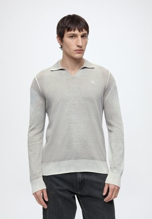 Giovane uomo che indossa una polo grigio chiaro a maniche lunghe e jeans scuri, in piedi davanti a uno sfondo bianco uniforme.