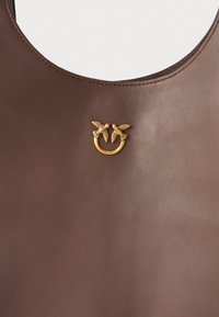 Bolsa de couro castanho com formato arredondado, textura suave e um detalhe em metal dourado em forma de círculo com elementos em folha.