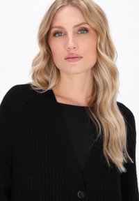 Cardigan nero lavorato a maglia con scollo a V e chiusura con un solo bottone. Il tessuto è testurizzato, con maniche lunghe e una vestibilità morbida.