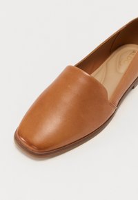 Brune skinn loafer med en glatt tekstur, firkantet tå og minimal søm. Flat såle med en subtil aksent nær hælen.