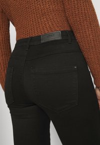 Svarta jeans med en slät yta, med två bakfickor och en läderetikett på midjan. Närbild från baksidan.