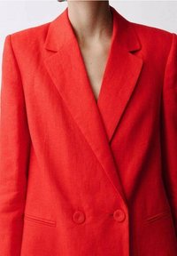 Blazer en lin rouge avec col cranté, devant croisé à double boutonnage avec boutons circulaires, et poches latérales. Texture lisse et coupe structurée.