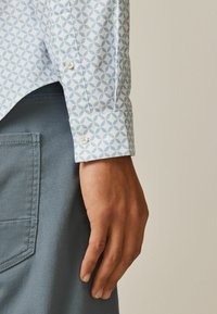 Lyseblå skjorte med mønster og små geometriske designs, med knapmanchetter. Kombineret med grå stof, der viser en baglomme.