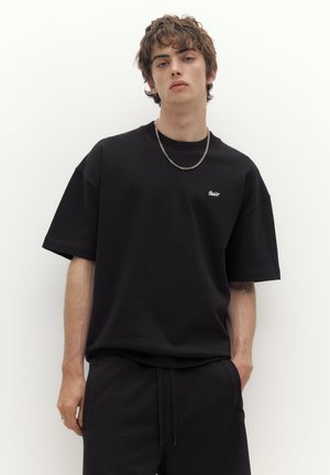 Jeune homme aux cheveux ébouriffés portant un T-shirt noir surdimensionné et un pantalon noir, debout devant un fond blanc uni.