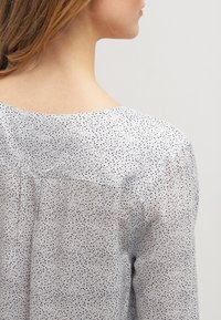 Lekka, przejrzysta bluzka wykonana z białej tkaniny w czarne plamki, z zaokrąglonym dekoltem i delikatnym zakładką z tyłu.