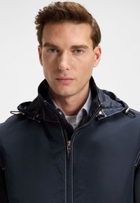 Tamsiai mėlyna vandeniui atspari striukė su kapišonu, priekyje su užtrauktuku ir sidabro spalvos detalėmis. Yra baltos siūlės ir laisvas kirpimas, matomas atvartas.