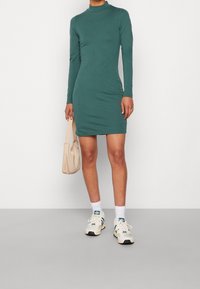 Zielona sukienka z długim rękawem, o wysokim dekolcie, dopasowanym kroju i miękkiej fakturze. Połączona z białymi skarpetkami, beżową torbą i trampkami.