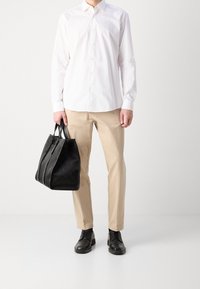 Camicia bianca con bottoni, pantaloni beige, scarpe eleganti nere in pelle e una grande borsa nera con superficie testurizzata, tenuta nella mano destra.
