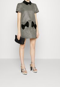 Svartvitmönstrad kort klänning med krage, med svarta sammetsrosetter på framfickorna. Kombinerad med silverfärgade tjockklackade sandaler.