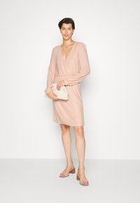 Vestido envolvente rosa pálido com mangas longas, tecido texturizado, decote em V profundo, cintura ajustada e silhueta fluida, acompanhado de uma pequena clutch.