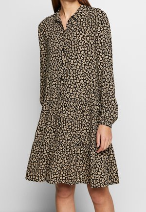 Frau trägt ein langärmliges Kleid mit Kragen, schwarz-beigem gepunktetem Muster, mit Stufenrock und Knopfleiste vorne.