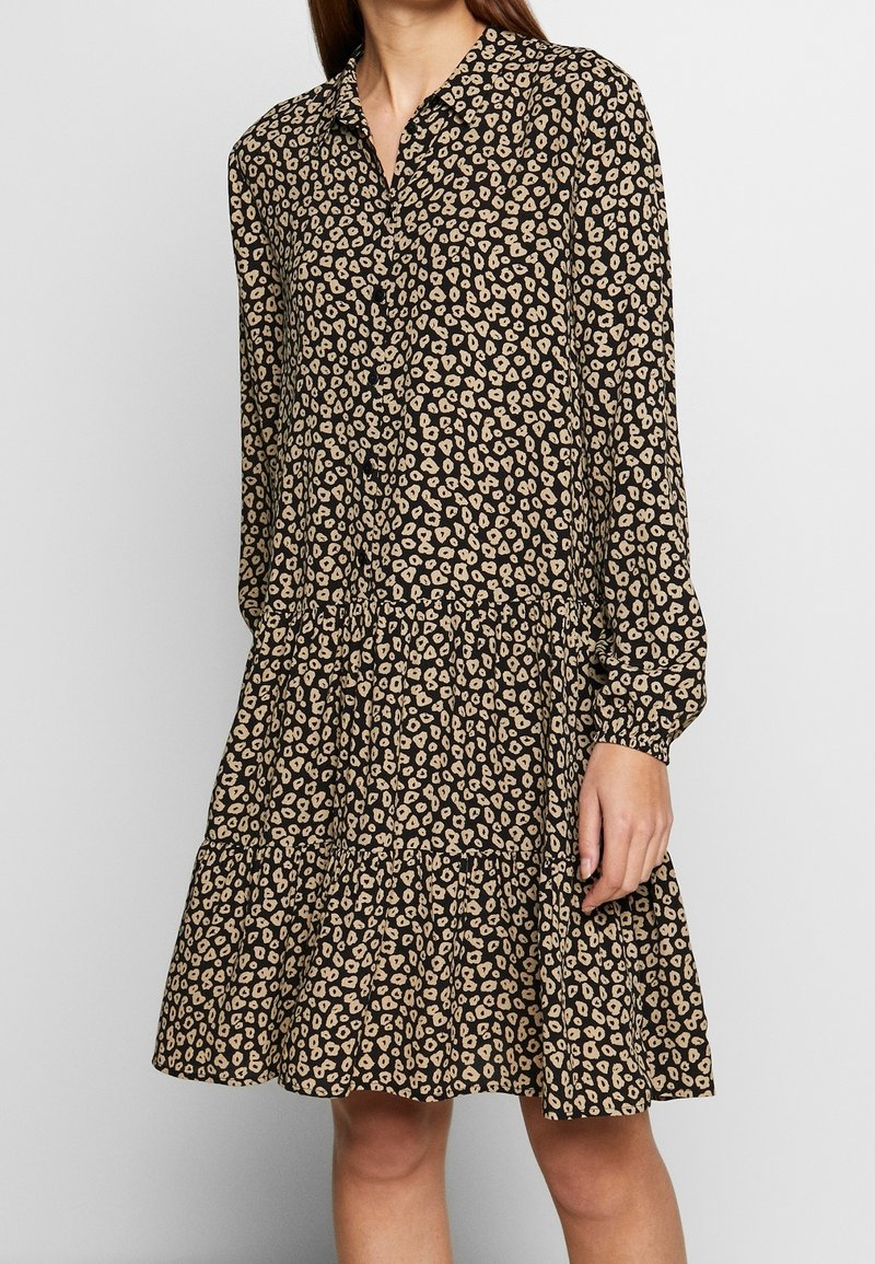 Femme portant une robe à manches longues avec col, à motif tacheté noir et beige, dotée d'une jupe à étages et d'un devant boutonné.