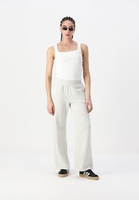 White tank top, grey sweatpants, black shoes with white stripes. Model standing with braided hair, minimal accessories, against a plain backdrop.