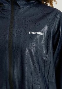 Vanntæt navy regnjakke med skinnende tekstur, der har en frontlynlås, sorte detaljer, og ordet "TRENTORN" i hvid på brystet.