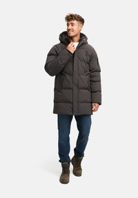 Giacca puffer isolante grigio scuro con cappuccio, caratterizzata da una chiusura con zip, due tasche laterali e polsini elasticizzati, abbinata a jeans blu e stivali marroni.