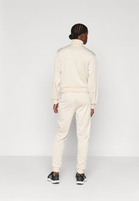 Beige träningsdräkt med hög krage och elastiska muddar, figursydd design, slät textur, kombinerad med svarta sportskor.