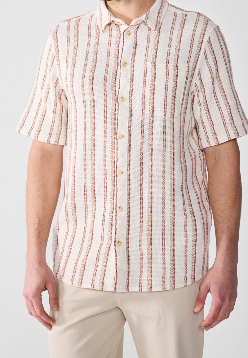 Homme portant une chemise blanche à manches courtes avec des rayures rouges verticales et des boutons beiges, associée à un pantalon beige clair, sur un fond uni.
