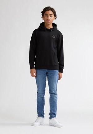 Sweatshirt noir à capuche avec une poche avant et un patch logo, associé à un jean denim bleu clair et des baskets blanches. Tenue simple et décontractée.