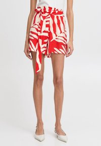 Žena v šortkách s abstraktním červenobílým vzorem, s látkovým pásem uvázaným v pase, doplněná bílými špičatými balerínami.