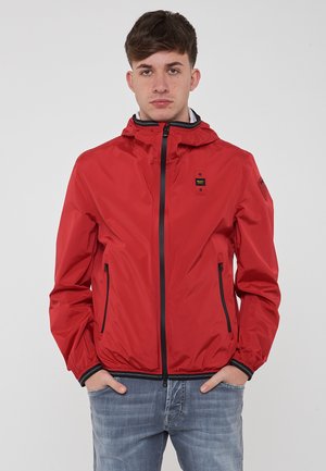 Giacca impermeabile rossa con zip frontale, cappuccio regolabile e due tasche laterali. Presenta dettagli neri e un logo sul petto.