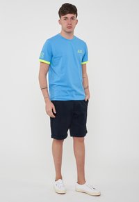 T-shirt di cotone azzurro chiaro con dettagli verdi, collo tondo, maniche corte e un logo sul petto. Abbinato a pantaloni corti scuri e scarpe bianche.