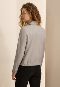 Grå bomberjakke med en glat tekstur, der har krave, frontlommer og elastisk talje. Stylet med sorte bukser.