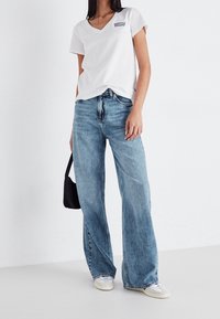 Valkoinen puuvillainen v-aukkoinen t-paita, jossa harmaa logo, yhdistetty vaaleansinisillä liehuleveillä farkuilla ja valkoisilla tennareilla, pitäen pientä mustaa laukkua.