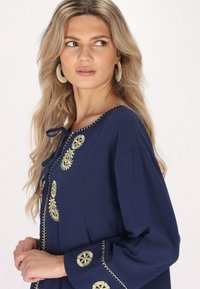 Tunic blu navy con ricami dorati, caratterizzata da un collo con laccetto, maniche lunghe e cuciture decorative lungo il colletto e le maniche.