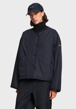 Femme portant une veste matelassée noire, un col roulé noir, un pantalon noir et une casquette noire avec un logo brodé, debout devant un fond uni.