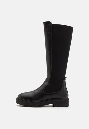 Melni ceļgala zābaki ar gludu ādas purngala daļu un gumijas zoli. Iekļauj stiepjamus auduma paneļus un cilpu augšējā daļā vieglai uzvilkšanai.